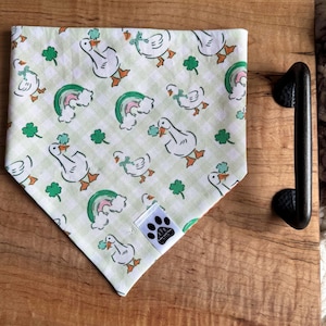 May include: A dog bandana with a light green gingham pattern featuring white ducks, green shamrocks, and rainbows. The bandana has a small tag with a paw print logo. The bandana is on a wooden tray.