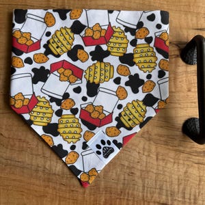 May include: A white dog bandana with a repeating pattern of chicken nuggets, french fries, and black cow spots. The bandana has a red border and a small tag with a paw print logo. The bandana is on a wooden tray.