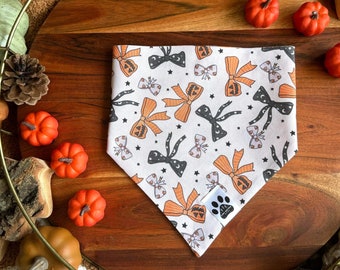 May include: A white bandana with a Halloween-themed print featuring orange and black bows, jack-o'-lanterns, and stars. The bandana is on a wooden tray with fall decorations.