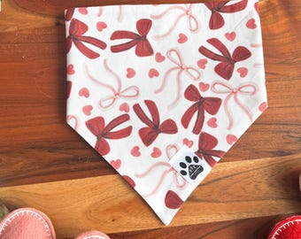 May include: A white pet bandana with a repeating pattern of red bows and pink hearts. The bandana is folded and placed on a wooden surface, surrounded by felt hearts and pink felt balls. A small tag with a paw print is visible.