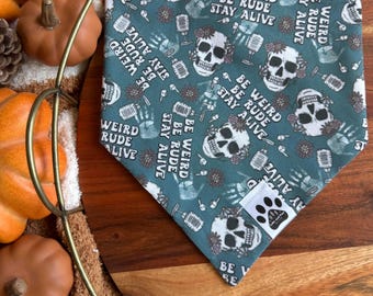 Op de afbeelding: Teal honden bandana met een herhalend patroon van suikerschedels, microfoons en de tekst "BE WEIRD, BE RUDE, STAY ALIVE." Inclusief een wit label met een pootafdruk. Perfect voor Halloween of dagelijks gebruik.