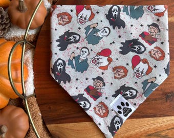 Op de afbeelding: Een lichtgekleurde hondenbandana met een herhaald patroon van horrorfilmkarakters, waaronder Michael Myers, Freddy Krueger en Pennywise. De bandana heeft een klein wit label met een pootafdruklogo.