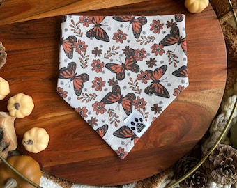 May include: A white bandana with a brown and orange butterfly pattern. The bandana has a small paw print logo in the bottom right corner.