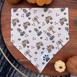 May include: A white bandana with a fall-themed print featuring scarecrows, pumpkins, and wagons. The bandana has a white paw print logo in the bottom right corner.