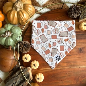 May include: A white bandana with a brown and orange pattern of books, coffee cups, and leaves. The bandana is on a wooden surface with small pumpkins around it.