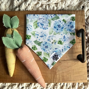 May include: A white bandana with a blue and green floral print. The bandana is on a wooden tray with two plush carrot toys.