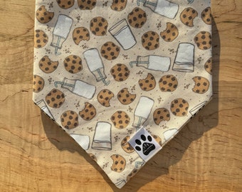 May include: A beige bandana with a repeated pattern of chocolate chip cookies and glasses of milk. The bandana has a small paw print logo in the bottom center.