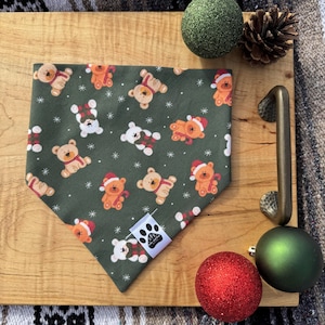 May include: A green bandana with a repeating pattern of brown and white teddy bears wearing Santa hats. The bandana is on a wooden surface with green and red Christmas ornaments.
