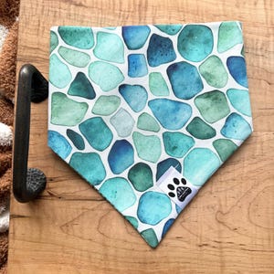May include: A blue and green patterned dog bandana with a white background. The bandana features a repeating pattern of irregular shapes in shades of blue and green. A small white tag with a paw print logo is sewn on the corner.