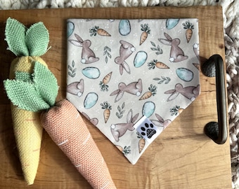 May include: A beige bandana with a repeating pattern of brown bunnies, blue eggs, and orange carrots. The bandana is on a wooden tray with two plush carrot toys.