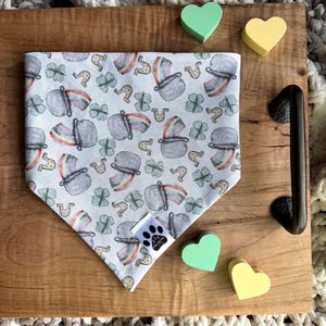 May include: A white bandana with a green, gold, and rainbow pot of gold pattern. The bandana is on a wooden surface with green and yellow heart-shaped objects.