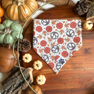 May include: A brown and tan bandana with a pattern of orange flowers, brown leaves, and white ghosts in mugs. The bandana is on a wooden surface with small pumpkins and a log.