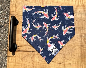 Puede incluir: Un pañuelo azul marino para mascotas con un estampado de peces koi en tonos rosa, amarillo y blanco. El pañuelo triangular tiene una pequeña etiqueta con un logotipo de huella de pata. Se muestra sobre una tabla de madera.