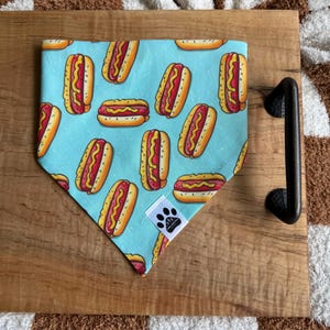 May include: A light blue dog bandana featuring a repeating pattern of cartoon hot dogs with yellow mustard and red ketchup. The bandana is triangular and has a small tag with a paw print logo. The bandana is on a wooden tray.