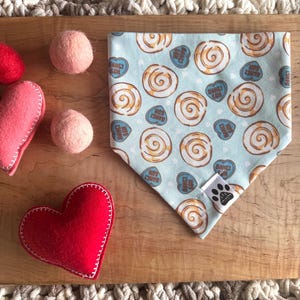 May include: A light blue dog bandana with a pattern of cinnamon rolls and hearts with the text "Hot Cinnamon Bun" on them. The bandana has a white paw print logo in the bottom right corner.