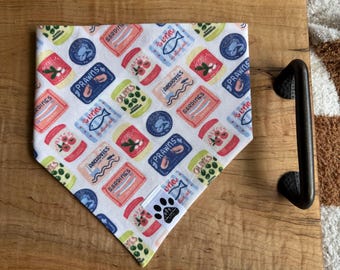 Puede incluir: Bandana blanca para perros con un colorido estampado de mariscos enlatados, incluyendo sardinas, anchoas, gambas y aceitunas. La bandana tiene una etiqueta con huella de pata y se muestra sobre una superficie de madera.
