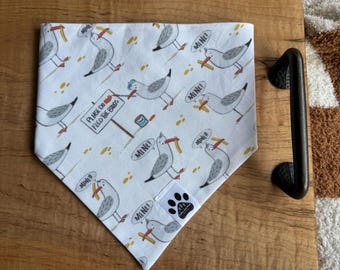 Puede incluir: Bandana blanca para perros con un patrón repetido de gaviotas de dibujos animados, cada una sosteniendo una patata frita y diciendo "MINE!". La bandana también presenta el texto "PLEASE DO NOT FEED THE BIRDS". Un logotipo de huella de pata es visible.