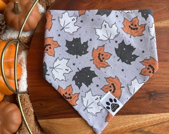 Op de afbeelding: Een grijze bandana voor honden met een herhalend patroon van witte, oranje pompoen- en donkergrijze esdoornbladeren. De bandana heeft een klein wit label met een pootafdruklogo. Op een houten oppervlak.
