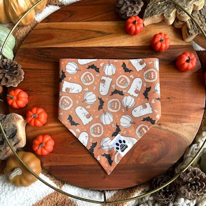 May include: An orange Halloween-themed dog bandana with a repeating pattern of white ghosts, pumpkins, and bats. The bandana is folded and displayed on a wooden tray with decorative pumpkins and pinecones.