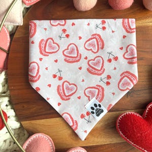 May include: A white dog bandana with a repeating pattern of pink heart-shaped cookies with the word "Love" and red cherries. The bandana has a tag with a paw print logo. Other felt hearts and pink balls are in the background.