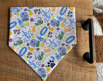 Puede incluir: Bandana blanca y azul para mascotas con un patrón repetido de ilustraciones de comida y bebida. El diseño incluye botellas de vino, copas, cangrejos, limones y pan. Se ve una pequeña etiqueta con un logotipo de huella de pata.