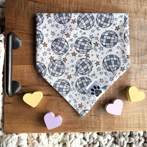 May include: White dog bandana with a disco ball pattern. The bandana has gold stars and confetti. The bandana is on a wooden surface with four small heart-shaped objects.