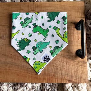 May include: White dog bandana with a St. Patrick's Day theme. The bandana features green dinosaurs wearing leprechaun hats and shamrocks. A small tag with paw prints is visible. The bandana is on a wooden tray.