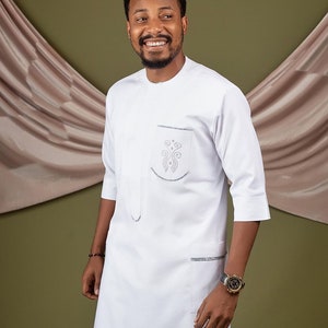 May include: A white long-sleeved shirt with a mandarin collar and a silver embroidered design on the chest pocket. The shirt has a single pocket on the left side and a second pocket on the right side.