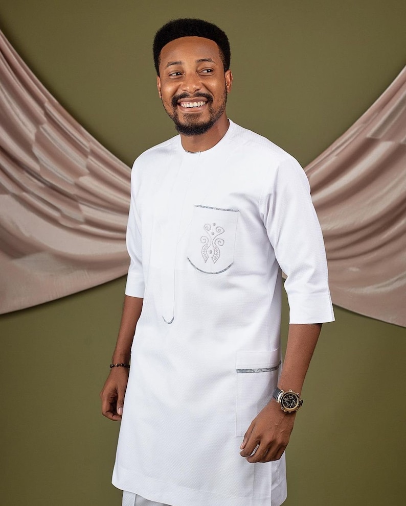 May include: A white long-sleeved shirt with a mandarin collar and a silver embroidered design on the chest pocket. The shirt has a single pocket on the left side and a second pocket on the right side.