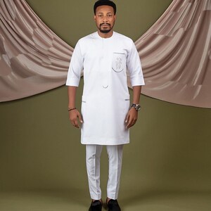 May include: A white long-sleeved shirt with a mandarin collar and a small embroidered design on the chest pocket. The shirt is paired with white pants and black shoes.