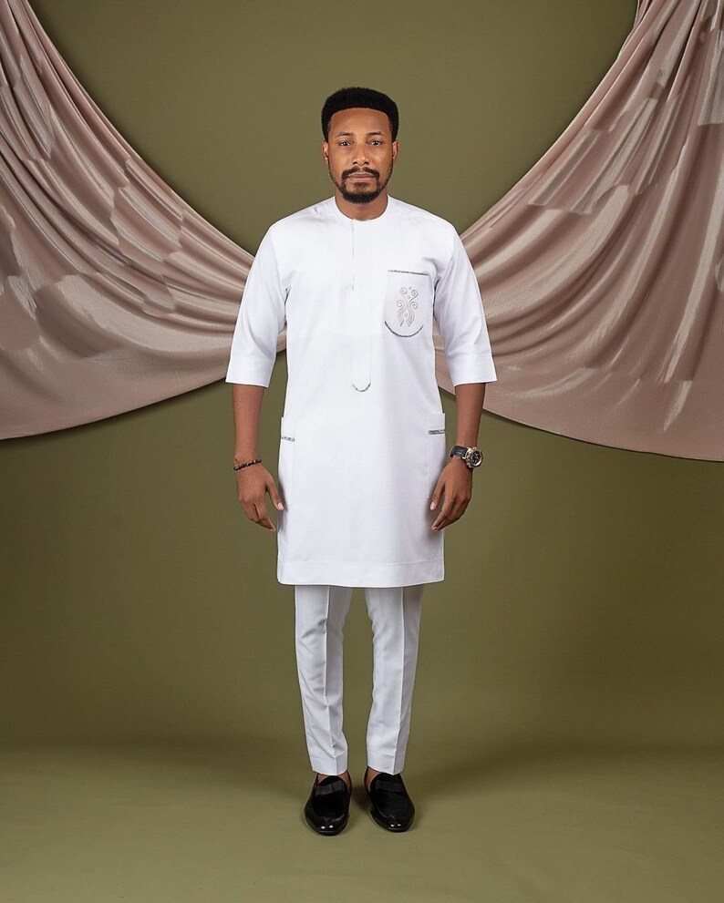 May include: A white long-sleeved shirt with a mandarin collar and a small embroidered design on the chest pocket. The shirt is paired with white pants and black shoes.