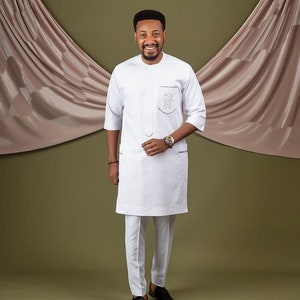 May include: A white long-sleeved shirt with a mandarin collar and a small embroidered design on the chest. The shirt is paired with white pants and black shoes.