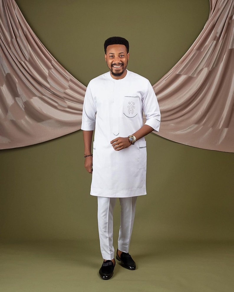 May include: A white long-sleeved shirt with a mandarin collar and a small embroidered design on the chest. The shirt is paired with white pants and black shoes.