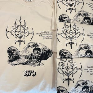 May include: A beige t-shirt with a black graphic print of a skull and crossbones design. The graphic includes the text "I can't lie to you about your chances, but you have my sympathies." and "1979".