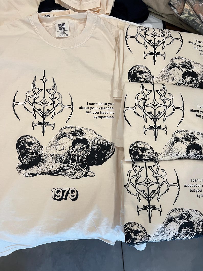 May include: A beige t-shirt with a black graphic print of a skull and crossbones design. The graphic includes the text "I can't lie to you about your chances, but you have my sympathies." and "1979".