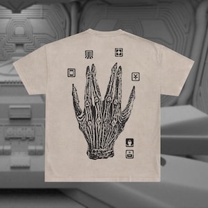 May include: A beige t-shirt with a black graphic print of a hand with multiple fingers. The hand is surrounded by small black squares with symbols inside.