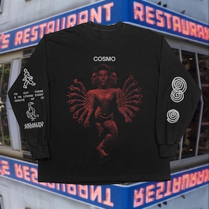 May include: Black long-sleeve shirt with a red illustration of a multi-armed figure and the word "COSMO". The left sleeve has white graphics and text, including "I'M OUT THERE & I'M LOVING EVERY MINUTE OF IT!". The right sleeve has white spiral designs.