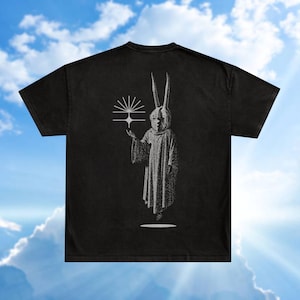 以下が含まれることがあります： 黒いTシャツ。白いイラストは、ウサギのようなマスクと角を持つ人物が幾何学的な形を持っている様子を描いています。デザインはTシャツの背面にあります。このTシャツは、色あせたヴィンテージの外観をしています。