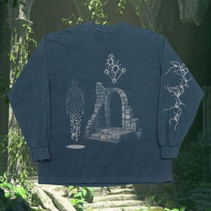 Può includere: Maglietta a maniche lunghe blu scuro con un design grafico. Il design include una figura punteggiata, una struttura ad arco in pietra e un sole astratto. La manica presenta un design intricato e fluido.