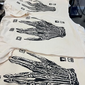 May include: Four white t-shirts with a black and white graphic print of a hand with intricate details. The hand is stylized and appears to be robotic or mechanical.