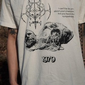 May include: A white t-shirt with a black graphic print of a skull and crossbones, a geometric design, and the text "I can't lie to you about your chances. but you have my sympathies. 1979".