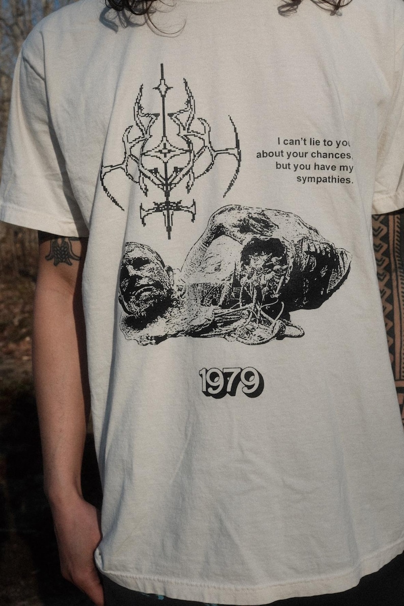 May include: A white t-shirt with a black graphic print of a skull and crossbones, a geometric design, and the text "I can't lie to you about your chances. but you have my sympathies. 1979".