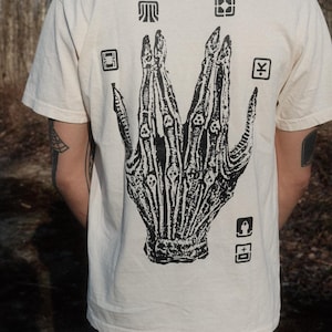May include: A white t-shirt with a black graphic of a skeletal hand with symbols around it. The symbols include a square, a yen sign, a lock, and a rectangle.