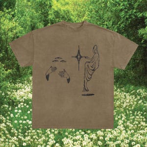 May include: A tan t-shirt with a black graphic design. The design features a figure, hands, and a starburst shape. The shirt has a crew neck and short sleeves. The background shows green foliage and white flowers, creating a natural setting.