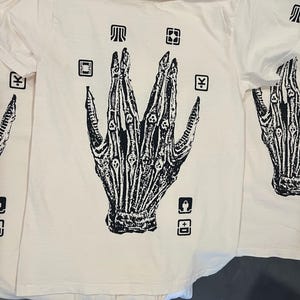 May include: A white t-shirt with a black graphic of a skeletal hand with multiple fingers. The hand is surrounded by small black squares with symbols inside.