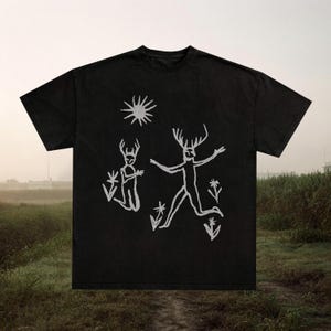 Puede incluir: Camiseta negra con un dibujo blanco de dos figuras con cuernos, un sol y flores. El diseño tiene una estética dibujada a mano. Es una prenda de vestir informal.