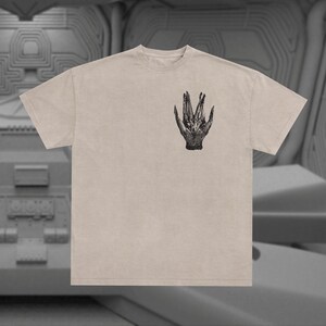 May include: A beige t-shirt with a black and white graphic print of a hand with three fingers extended. The hand is reminiscent of the alien creature from the movie Alien.