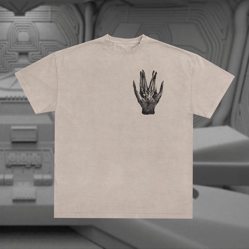 May include: A beige t-shirt with a black and white graphic print of a hand with three fingers extended. The hand is reminiscent of the alien creature from the movie Alien.
