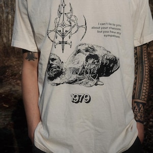 May include: A white t-shirt with a black graphic print of a skull and a symbol. The text on the shirt reads "I can't lie to you about your chances, but you have my sympathies. 1979"