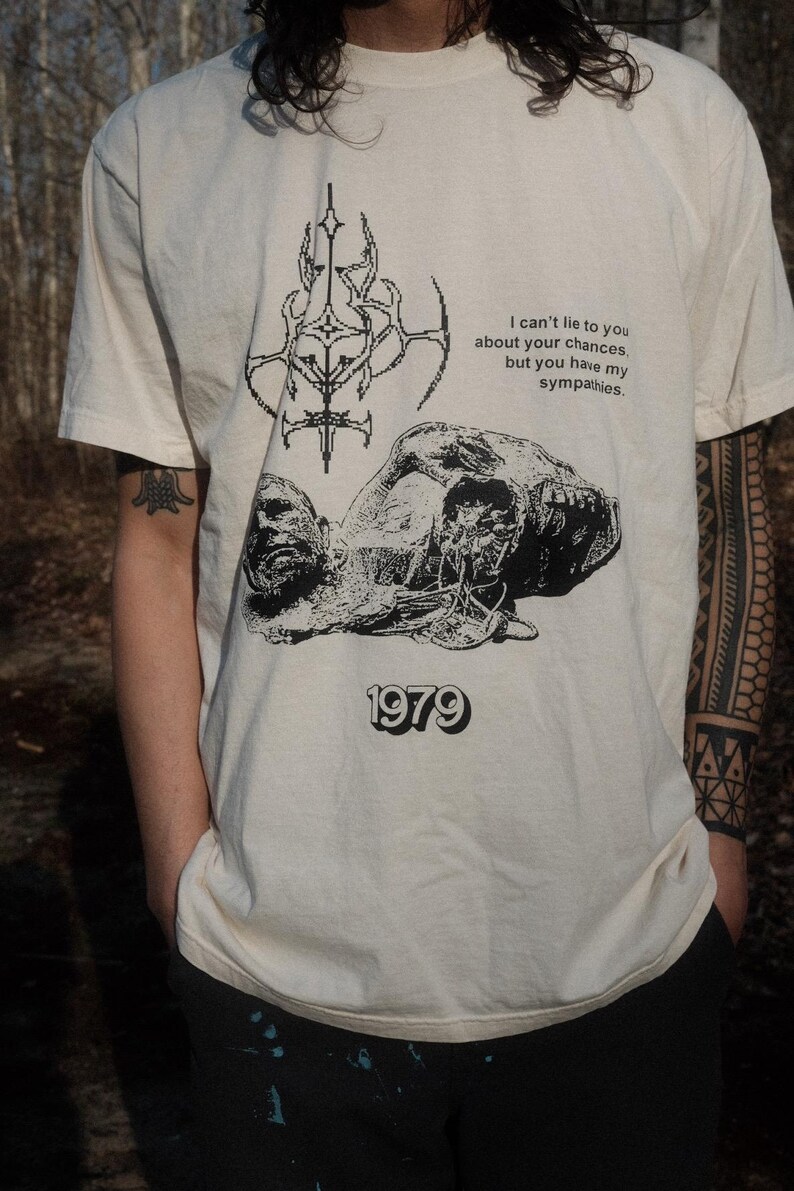 May include: A white t-shirt with a black graphic print of a skull and a symbol. The text on the shirt reads "I can't lie to you about your chances, but you have my sympathies. 1979"